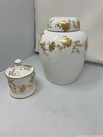 Set of two white fine bone china ginger jars with gold floral decoration, lids on, shown on gray surface.