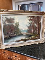 Framed oil painting on canvas with a landscape featuring water, trees, and distant mountains.