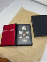 Top view of the opened black presentation box with red satin interior showing seven commemorative 1986 Canadian coins and accompanying outer black lid box.