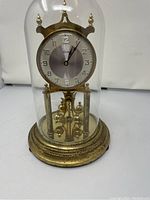 Full front view of vintage Kieninger & Obergfell brass clock under glass dome showing clock face, base and pendulum.