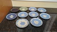 Eleven vintage Wedgwood plates laid out on a dark counter surface, mostly blue with one green and one dark plate, all with central white jasper relief decoration.