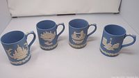 Four Wedgwood blue jasperware mugs each with a white relief landmark design, displayed in a row showing all four different scenes.