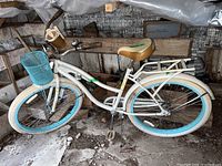 Full side view of white Huffy Nel Lusso bicycle with blue and white tires, front basket, cup holder, and tan saddle. Showing entire bike from left side.