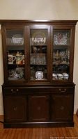 Front view of dark wood china cabinet showing three glass doors on upper section with displayed glassware and wood lower section with doors and drawer.
