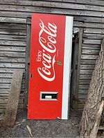 Full view of large red Coca Cola door/sign with white logo, peeling paint, scratches, metal bottle opener, and weathered condition leaning against a wooden structure outdoors.