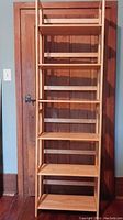 Collapsible wooden bookcase standing upright showing all shelves, light-colored wood with seven shelves, against closed wooden door.