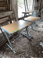 Two adjustable desks with silver metal X-frames and light wood tops in barn environment, showing surface scratches and dirt.