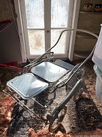 Vintage metal doll carriage with folding frame, blue metal seats, metal wheels, and a curved metal handle.
