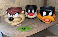 Three ceramic Looney Tunes character mugs displayed on a wooden surface, showing Taz, Sylvester, and Daffy Duck faces.
