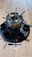 View of the stainless steel fondue pot with six fondue forks inserted and six black ceramic bowls arranged on a black tray.