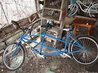 Side view of the blue Genesis tandem bicycle stored in a barn showing flat tires and the bike frame with two seats and two handlebars.