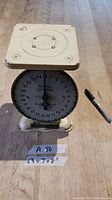 Front angle photo showing entire scale with square weighing platform, dial face visible, placed on wooden floor, pen for size reference.