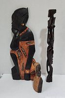 Three African style wooden decor pieces displayed together against a white background, showing a painted seated woman figure, a standing carved dark wooden statue, and a small carved bust.