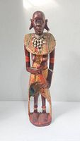 Front view of the wooden African statue showing full figure, beaded necklace, painted dress pattern, and visible cracks.