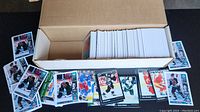 Top view of partially filled cardboard box with a partial collection of hockey cards both inside and laid out around the box showing various players, teams, and card designs.