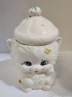 Front view of white ceramic cat cookie jar with lid showing blue eyes, yellow flower decorations, and details in fur texture.