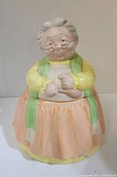 Front view of ceramic cookie jar shaped like a grandmother with glasses, wearing yellow shirt, green scarf, and peach skirt.