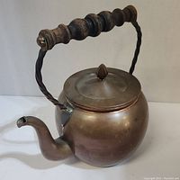 Side angle view of the copper kettle showing round copper body, lid with pointed knob, curved spout, and twisted metal handle with wooden grips.