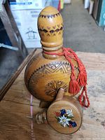 Wooden flask showing carved geometric designs with red tassel, paired with a small wooden jigger with colorful floral painting.