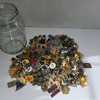 Empty glass jar and loose collectible pins spread out with various designs and colors, mainly gold and silver metal with enamel.