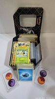 Black Pokemon-themed metal tin filled with assorted Pokemon trading cards and energy cards displayed outside in clear plastic holders.