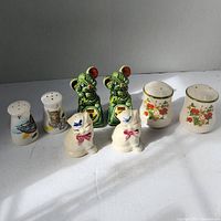 Photo showing all four sets of ceramic salt and pepper shakers arranged in a row on a gray background, including cat, mouse, bird/barrel, and floral designs