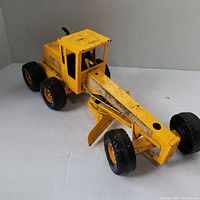 Yellow metal Nylint toy grader with an operator cab, four black wheels, and a blade attachment underneath the long arm extending from front to middle of grader. Paint shows visible wear and chipping.
