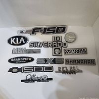 Photo showing 14 assorted vehicle emblems and badges including XL F-150, Kia, Chevrolet 1500, Silverado, Hyundai dealer badge, and others.