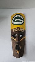 Front view of tall wooden mask carved with facial features, painted with Green Bay Packers logo and team name