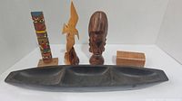 Five wood decor items displayed together in a white photo box: a multicolored totem pole, bird carving, mask sculpture, rectangular block, and black canoe tray.