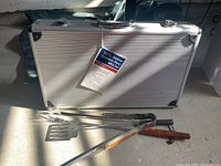 Closed aluminum storage case labeled '24 Pc Deluxe Stainless Steel BBQ Set 480 steel' alongside three loose tools - spatula, tongs, and fork with wooden handles.