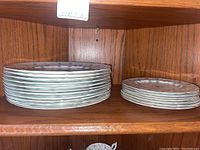 Stack of larger dinner plates and smaller luncheon plates on wooden shelf.