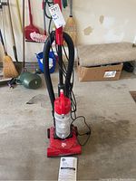Full view of red Dirt Devil Quick-Lite upright vacuum in garage with hose and power cord, manual on ground.