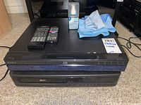 Sony BDP-S300 Blu-Ray disc player with remote and Zenith VRJ415 VHS player under it, screen cleaner and cleaning cloths on top, black electronic devices stacked on carpeted floor.