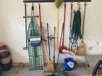 View of garage wall-mounted bar holding multiple garden tools such as brooms, dustpan, garden hose, shovel, bucket, and ironing board leaning against the wall.