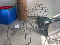 Two metal plant stands shaped like chairs, sitting on a concrete floor with sunlight casting shadows.