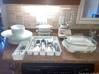 Wide shot of all items on countertop showing plates, bowls, cutlery trays, casserole dishes, and blender.