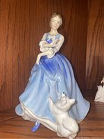 Porcelain figurine of a woman in a blue dress holding a child with a white dog at feet, on wooden background.