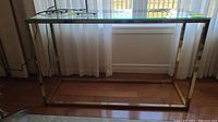 Wide shot of brass framed sofa table with clear glass top showing full piece in front of window and curtain.