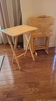 One wooden folding tray table set up, with three folded tray tables stacked behind it on wood floor.