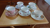 Five teacups and saucers arranged on a small wooden table, showing various floral patterns and gold trim detail.
