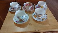Five vintage teacups and saucers displayed on wooden table, floral designs visible in various colors and gold edging