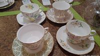 Photo showing four different teacups with matching saucers arranged in a group displaying floral and ornate patterns in various colors including pale green, pink, white, and detailed botanicals.