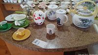 Overview of multiple teapots, mugs, cups including distinctive Villeroy & Boch kettle on center right, Sadler teapot center left with butterflies, Indigo set green and white cups, and small demitasses