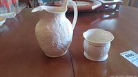 Photo showing Wedgwood pitcher and Belleek sugar bowl together on wooden table, pitcher with handle and floral grapevine design, sugar bowl smaller with reliefs.