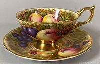 Side view of teacup positioned on saucer showing full fruit motif and gold handle
