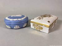 Two porcelain trinket boxes side by side: Wedgwood blue Jasperware scalloped box with white reliefs and a white rectangular Porcelaine De France box with gold details.
