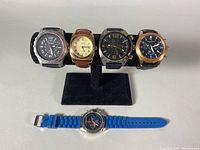 Front view of all five men's wristwatches on display stand and one watch laid flat showing blue rubber strap