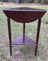 Front view of demi-lune triple drop leaf wooden side table standing on grass, showing curved half-moon drop leaf tabletop and lower triangular shelf with three legs.