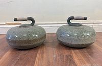 Two vintage BWK curling stones shown side by side, displaying front and handle details on wood floor